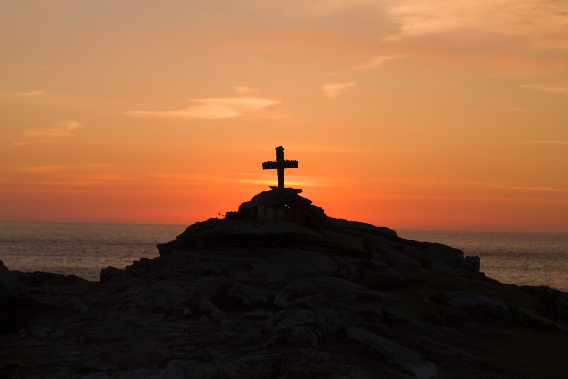 Backlit picture of a cross on top of a hill in front of a sunset.